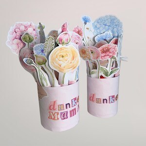 Puede incluir: Dos ramos de flores de papel rosa con el texto "danke Mama". Los ramos presentan una variedad de flores de papel coloridas, incluyendo hortensias, rosas y otras flores. Las flores est&aacute;n dispuestas en recipientes rosas cil&iacute;ndricos.