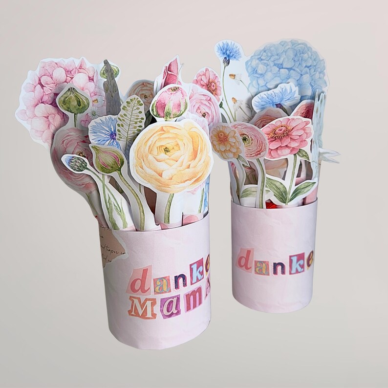 Puede incluir: Dos ramos de flores de papel rosa con el texto "danke Mama". Los ramos presentan una variedad de flores de papel coloridas, incluyendo hortensias, rosas y otras flores. Las flores est&aacute;n dispuestas en recipientes rosas cil&iacute;ndricos.
