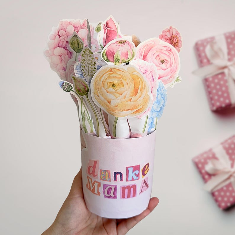 Puede incluir: Un jarr&oacute;n de flores de papel rosa con una variedad de flores de papel en tonos rosa, amarillo y azul. El jarr&oacute;n tiene las palabras "danke Mama" en letras coloridas. Dos regalos envueltos con lunares rosas est&aacute;n en el fondo.