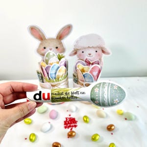 Könnte beinhalten: Osterdekorationen mit einem Hasen und einem Lamm, die Ostereier halten. Ein weißer Löffel mit Eierdesign und dem Text "du machst die Welt ein bisschen heller" wird in einer Hand gehalten. Bunte Süßigkeiten sind verstreut.