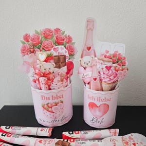 May include: Two pink Valentine's Day gift boxes with floral and teddy bear decorations. One box says "Du bist wunderbar" and the other says "Ich liebe Dich". Chocolate bars are at the bottom.