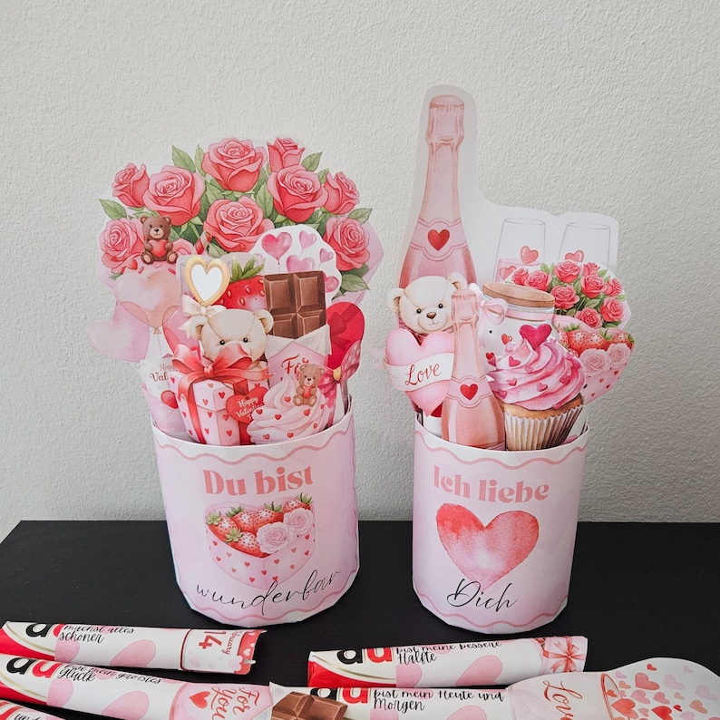 K&ouml;nnte beinhalten: Zwei rosa Valentinstags-Geschenkboxen mit Blumen- und Teddyb&auml;r-Dekorationen. Auf einer Box steht "Du bist wunderbar" und auf der anderen "Ich liebe Dich". Schokoriegel sind unten.
