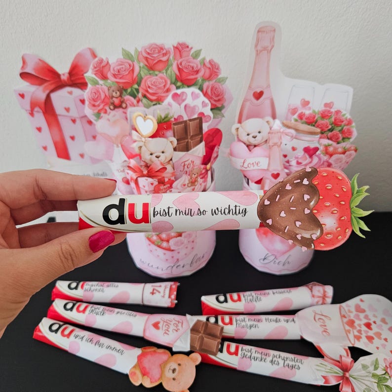 K&ouml;nnte beinhalten: Eine Valentinstags-Arrangement mit dekorativen Schokoriegeln, einer schokoladen&uuml;berzogenen Erdbeere und Blumenarrangements. Die Riegel tragen das Wort "du" und den Satz "bist mir so wichtig". Weitere Riegel sind sichtbar.