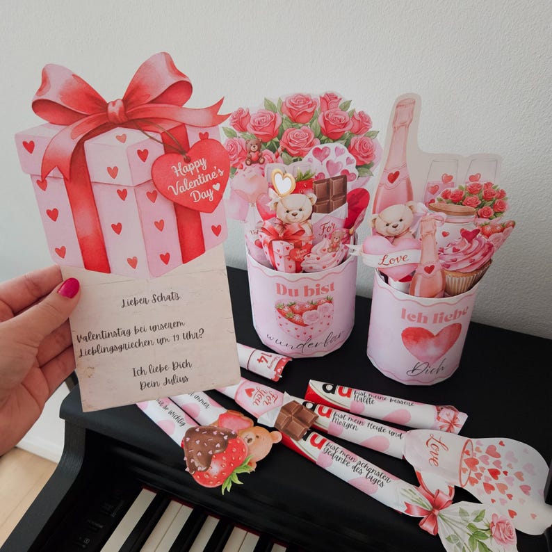 K&ouml;nnte beinhalten: Dekorationen zum Valentinstag. Enth&auml;lt eine Geschenkbox-Karte mit Schleife, Herzakzenten und dem Text "Happy Valentine's Day". Enth&auml;lt auch dekorative Becher mit Teddyb&auml;ren, Rosen und Schokolade sowie Schokoriegel.