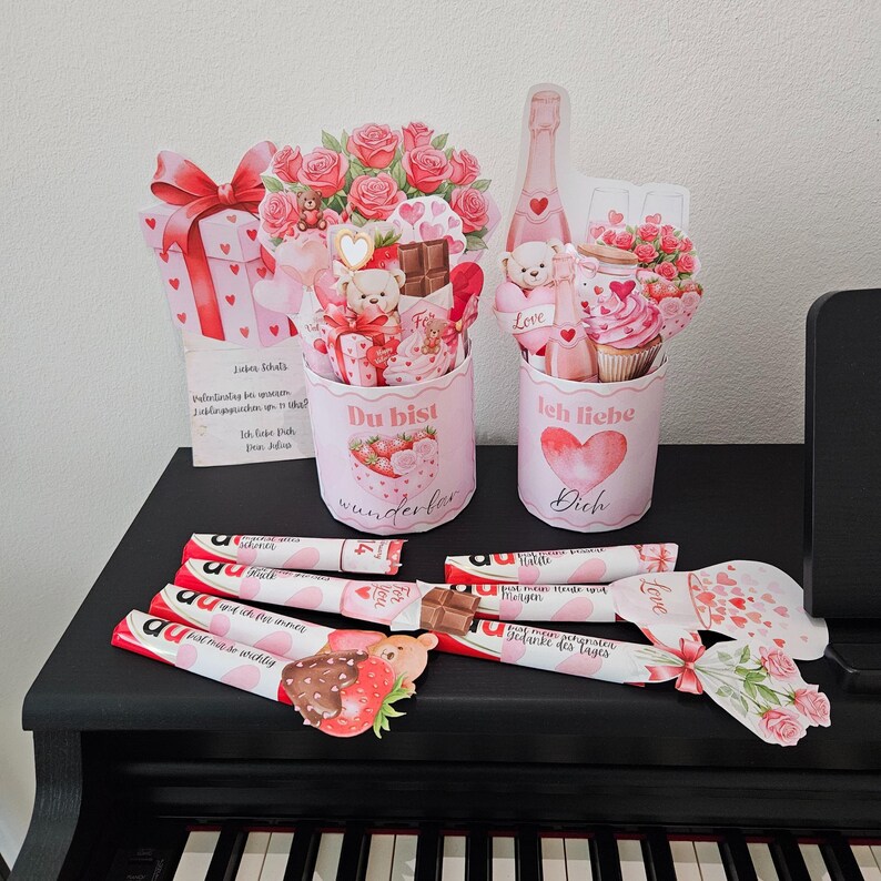 K&ouml;nnte beinhalten: Valentinstag-Geschenke und Dekorationen. Das Bild zeigt rosa Geschenkboxen, Papierstr&auml;u&szlig;e mit Rosen und Schokoriegel. Die Worte "Du bist wunderbar" und "Ich liebe Dich" sind auf den Geschenken zu sehen.