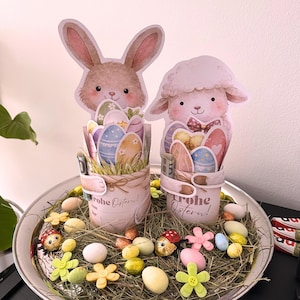 以下が含まれることがあります： イースターをテーマにした飾り。ウサギと子羊のフィギュアが、それぞれカラフルなイースターエッグのバスケットを持っています。銀色のトレイに、干し草、花、お菓子が飾られています。「Frohe Ostern!」の文字がバスケットに表示されています。