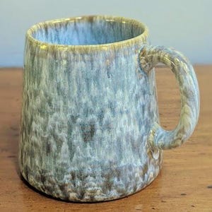 May include: A ceramic mug featuring a reactive glaze with a blend of muted blue, grey, and earthy brown tones. The mug has a cylindrical body with a slightly tapered base and a sturdy, curved handle on the side, showcasing a smooth, glossy finish.
