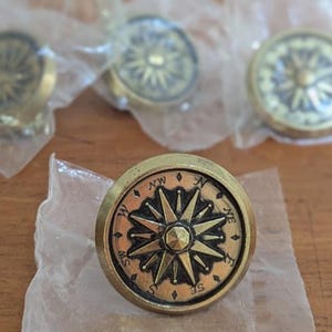 May include: Four round metal buttons featuring a raised compass rose design in a brass-toned finish. Each button is individually wrapped in a clear plastic bag, showcasing the detailed directional markings and central star motif on the face.