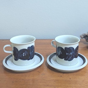 May include: Two off-white ceramic coffee cups with matching saucers, each featuring a dark blue floral pattern around the center. The saucers have a dark blue circular band along the rim, and the cups have a simple handle on the side.