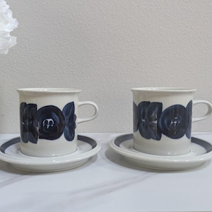 May include: Two off-white ceramic coffee cups with matching saucers, each featuring a dark blue painted floral pattern around the exterior. The cups have a cylindrical shape with a side handle, and the saucers have a dark blue circular rim.