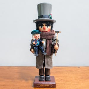 May include: A wooden nutcracker figurine depicting Bob Cratchit wearing a grey top hat, a brown scarf, and a long coat. The figure holds a small doll in one arm and a wooden cane in the other. It stands on a rectangular base labeled Bob Cratchit.