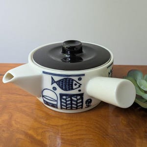 May include: A white ceramic teapot with a dark brown lid and a side handle. The body features a blue graphic pattern depicting a fish, a bucket, and geometric shapes. The teapot has a short, pointed spout and a cylindrical handle extending from the side.