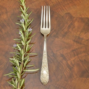 May include: A silver fork with an ornate handle rests on a dark wood surface next to a sprig of rosemary. The fork's tines are straight and parallel. The rosemary has green leaves and small purple flowers.