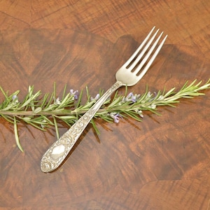 May include: A silver fork with an ornate floral design on the handle rests on a sprig of rosemary with small purple flowers. The fork is positioned diagonally against a dark brown, wood-grain surface.