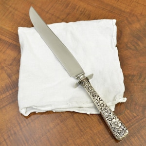 May include: A silver carving knife with a floral patterned handle rests on a white linen cloth. The knife's blade is long and shiny, reflecting the light. The handle features detailed floral engravings, adding a touch of elegance to the utensil.