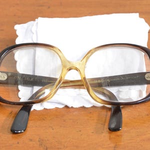 May include: Vintage eyeglasses with a brown and tan gradient frame and black temples. The glasses are resting on a white, scalloped-edge cloth. The lenses are clear. The frame has the text "CN 1621" and other text.