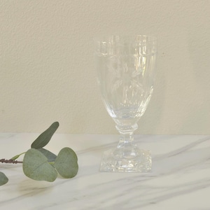 May include: A clear, etched glass goblet with a square base and a detailed floral design. The goblet has a flared top and a stem with a decorative element. The glass is set on a white marble surface.