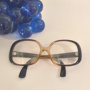 May include: A pair of vintage eyeglasses with a brown and black frame. The lenses are clear, and the frame has a rounded shape. The glasses are set against a white background, with a cluster of blue glass beads in the upper left corner.