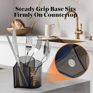 K&ouml;nnte beinhalten: Ein klarer, geteilter Utensilienhalter mit Edelstahlgabeln, L&ouml;ffeln und einem Messer. Der Halter tr&auml;gt den Text "Steady Grip Base Sits Firmly On Countertop" und ein goldenes Logo. Der Halter steht auf einer wei&szlig;en Arbeitsplatte.