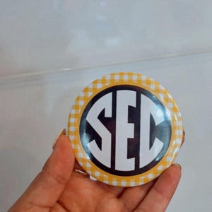 May include: A round button with a yellow and white gingham pattern border. The center features the letters "SEC" in a black and white circular design. The button is held in a hand.