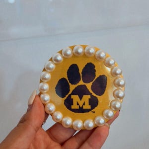 May include: A round, yellow decorative item featuring a black paw print with the letter "M" and surrounded by white pearls. The item is a University of Michigan design.