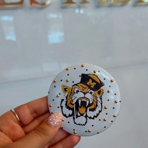 May include: A white round button with a tiger graphic wearing a black and gold hat. The button is decorated with small gold studs around the edge. The word "ESTABL" is visible in the background.