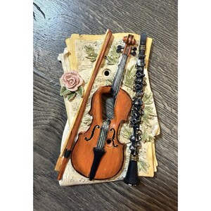 May include: A decorative light switch plate featuring a relief design of a brown violin, a black clarinet, and a bow resting on a background of cream-colored sheet music with green leaves and a single pink rose accenting the left side.