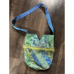 May include: A handmade shoulder bag with a blue strap and a body featuring a mix of green and blue fabric. The bag has a unique shape and is adorned with a fringe of yellow-green material. The bag is placed on a wooden surface.