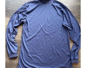 May include: A long-sleeved, periwinkle turtleneck top. The top features a high, folded collar and long sleeves with cuffs. The fabric looks soft and drapes loosely. The top is laid flat on a wooden surface.