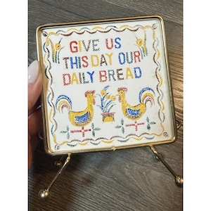 Puede incluir: Un salvamanteles rectangular vintage con marco y soporte de metal dorado. La baldosa de cerámica blanca presenta el texto bordado "GIVE US THIS DAY OUR DAILY BREAD" sobre dos gallos amarillos y una planta en maceta.