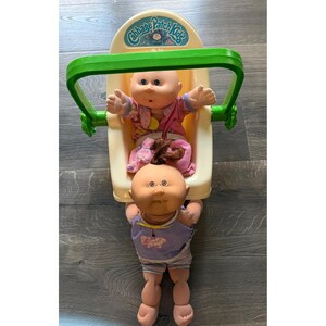 以下が含まれることがあります： 2体のキャベッジパッチキッズ人形。1体は緑色のハンドルとキャベッジパッチキッズのロゴが付いたクリーム色のプラスチック製キャリアに座っており、ピンクの柄の服を着ています。もう1体は前に立っており、紫色のトップスとストライプのショートパンツを着用しています。