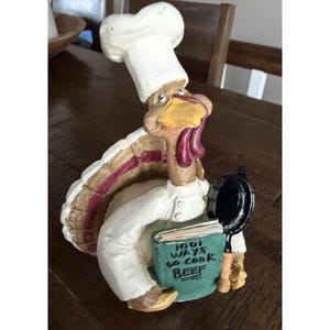 May include: A turkey figurine dressed as a chef in a white hat and jacket, holding a green book titled 1001 WAYS TO COOK BEEF and a small black frying pan. The turkey has brown feathers with red accents and a yellow beak with a red wattle.
