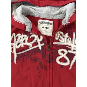 May include: Red zip-up hoodie with the Aeropostale brand name in white and gray lettering. The hoodie has a gray hood lining and the size tag reads XL/TG. The design includes the number 87 and other graphics.