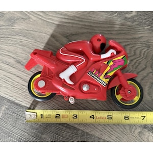May include: A red toy motorcycle with a matching rider figure. The motorcycle has yellow wheels and the text "SUPERMOTOR" on the side. The image includes a measuring tape showing the toy's length.