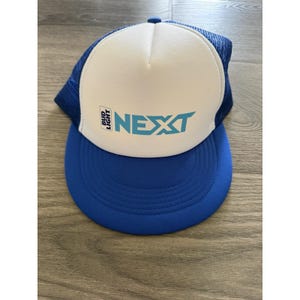 Op de afbeelding: Een blauw-witte truckerpet met een schuimrubberen voorpaneel waarop de tekst Bud Light NEXT in lichtblauw en zwart is gedrukt. De pet heeft een effen blauwe klep en een blauw mesh achterpaneel met een knoopje boven op de kroon.