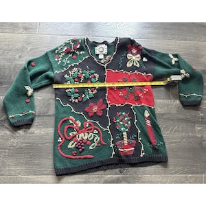 Könnte beinhalten: Ein langärmeliger V-Ausschnitt-Pullover in Dunkelgrün, Schwarz und Rot mit gestickten Weihnachtsmotiven wie Kränzen, Weihnachtssternen, einer Kerze und einem Formgehölz. Ein gelbes Maßband zeigt eine Breite von etwa 58,5 Zentimetern.