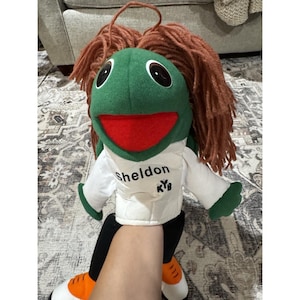 May include: A plush puppet with a green head, large black eyes, and a red mouth. It has brown yarn hair and wears a white shirt with the name "Sheldon" and "KYB" printed on it. The puppet's arms and legs are black and white.