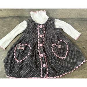 May include: A grey polka-dot dress with pink heart-shaped pockets and trim, worn over a white turtleneck top. The dress has pink heart-shaped buttons and bows. The dress is designed for a child.