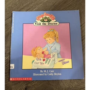 May include: A Cabbage Patch Kids Visit the Doctor paperback book by M.J. Carr and illustrated by Cathy Beylon. The cover features a blonde child using a stethoscope on a doll, with a medical kit on a table, set against a light blue background.