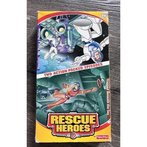 Puede incluir: Una cinta VHS vintage de "Rescue Heroes" con dos episodios llenos de acción: "Houston, We Have a Problem" y "Trapped Beneath the Sea". La portada presenta ilustraciones de dibujos animados de astronautas y buzos en colores vibrantes, con el logo de Fisher-Price.