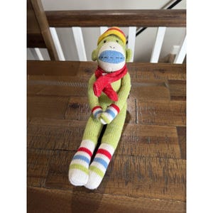 May include: A knitted sock monkey toy featuring a light green body, a red scarf, and a yellow and red cap. The monkey has a cream-coloured face with a blue stitched mouth, and its arms and legs have striped cuffs in red, white, and light blue colours.