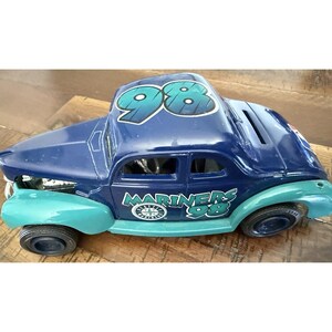 May include: A blue and teal Mariners '98 car-shaped money box. The car features the Mariners logo and the number 98 on the roof and side. It has black tyres and sits on a wooden surface.