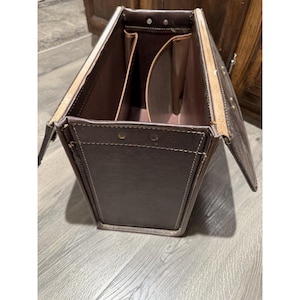 May include: Open brown leather briefcase with a rectangular shape. The briefcase has a light brown interior with several compartments and is secured with metal rivets. The edges are reinforced with lighter-colored leather and stitching.