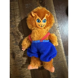 May include: A vintage plush lion doll with orange fur, blue eyes, and a smiling face. The doll wears blue shorts with red braces. The doll's arms and feet are a peach colour.
