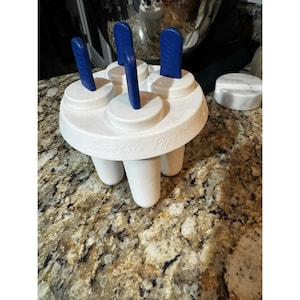 May include: White "Smoothie Pops" ice pop maker with blue handles. The maker has a circular design with four molds and a separate lid. The base has three legs. The text "Smoothie Pops" is printed on the side.