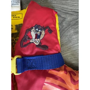 Puede incluir: Un chaleco salvavidas rojo y amarillo para niños con una imagen bordada del personaje de dibujos animados Taz. El chaleco tiene una correa azul con una hebilla amarilla y está diseñado para niños de 13,6-22,7 kg.