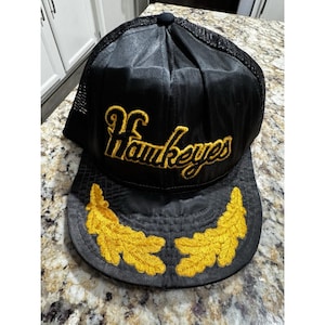 Puede incluir: Gorra de camionero negra con la palabra "Hawkeyes" bordada en oro. La gorra tiene una parte trasera de malla y una visera curva con bordado de hojas doradas. Un accesorio deportivo clásico.