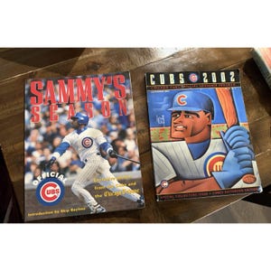 May include: Two Chicago Cubs baseball publications. The left book is titled Sammy's Season with a photo of a player in a white pinstripe uniform. The right magazine is titled Cubs 2002 Chicago Cubs Official Souvenir Program featuring a Corey Patterson illustration.