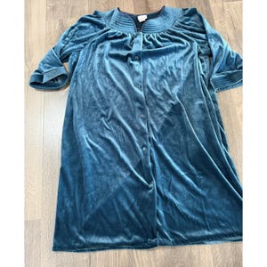 May include: A teal-colored, button-down robe made of soft velvet fabric. The robe has a round neckline with a pleated design and three-quarter sleeves. The robe is knee-length and has a relaxed fit.