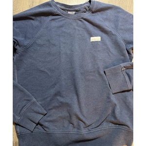 May include: A navy blue sweatshirt with a crew neck and long sleeves. The garment features a small rectangular brand patch on the chest. The cuffs and hem are ribbed.